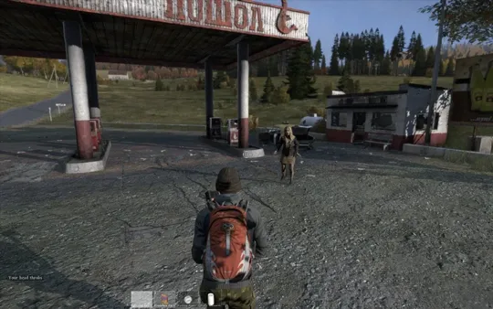 A DayZ survivor with a red hiking backpack approaches a dilapidated, overgrown gas station in a rural landscape.