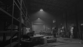 Black and white of a warehouse with a truck near a closed bay door. There's what looks to be a zombie shambling away from the camera.