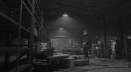 Black and white of a warehouse with a truck near a closed bay door. There's what looks to be a zombie shambling away from the camera.