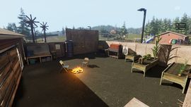 Screenshot from game showing a couple of folding chairs around a campfire with makeshift raised food gardens, a shooting target, and various other tools. Everything is surrounded in plywood with giant plywood doors. Set in a field surrounded by pine trees.
